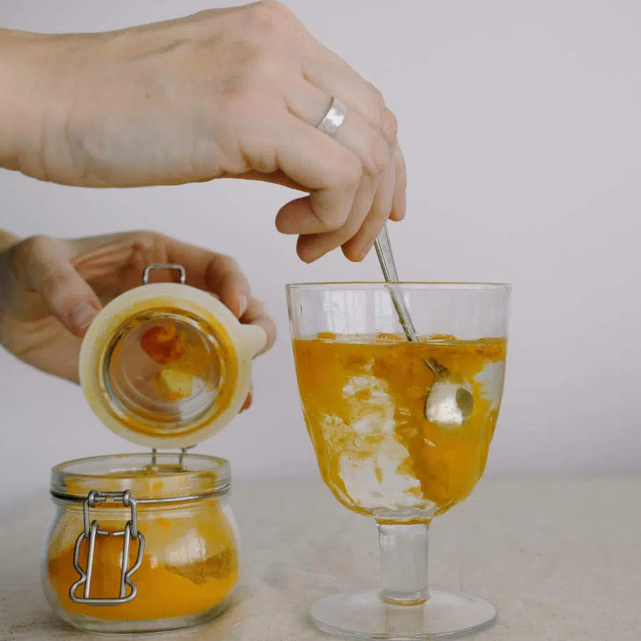 Die rechte Hand einer Frau rührt einen Teelöffel Kurkumapulver in ein Glas mit Wasser. Ihre linke Hand hält ein Vorratsglas mit Kurkumapulver am Deckel geöffnet.