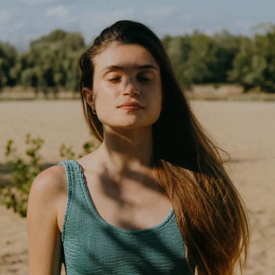 Eine junge Frau mit langen braunen Haaren steht inmitten eines Feldes. Sie hat die Augen geschlossen und steht halb in der Sonne und halb im Schatten einiger Baumäste.