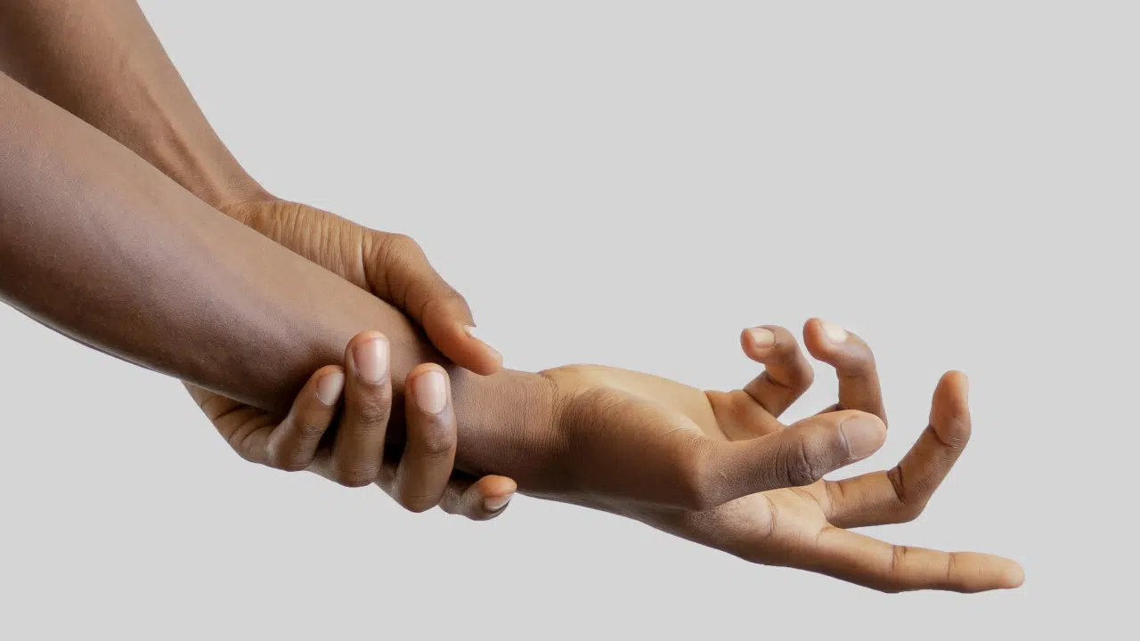 Zwei nackte Hände sind vor weißem Hintergrund. Die linke Hand umfasst das rechte Handgelenk. Die Finger der rechten Hand sind aufgefächert und gekrümmt.