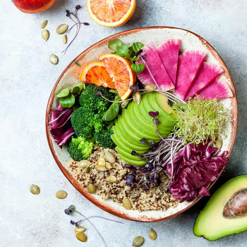 Eine bunte Bowl voller gesunder Lebensmittel wie Avocado, Orangenscheiben, Quinoa, Sprossen und Brokkoli.