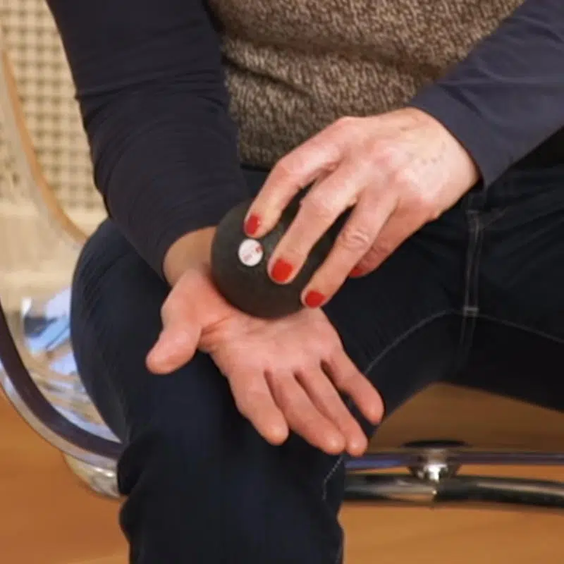 Eine Frau hat ihre rechte Hand auf dem Oberschenkel abgelegt und massiert ihre Handfläche und den Daumenballen mit einer kleinen Faszienkugel.