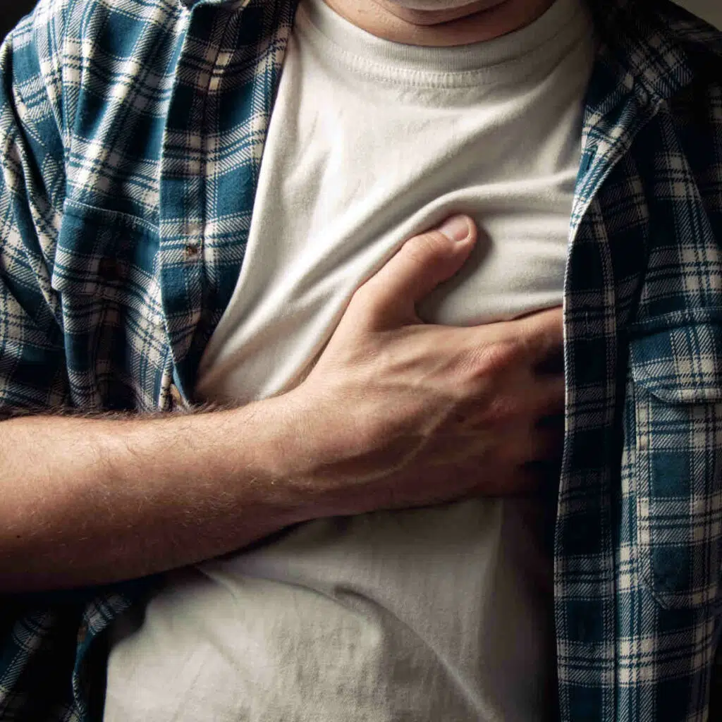 Ein Man im Karo-Hemd und einem beigen Shirt darunter greift sich mit der rechten Hand an die Brust.
