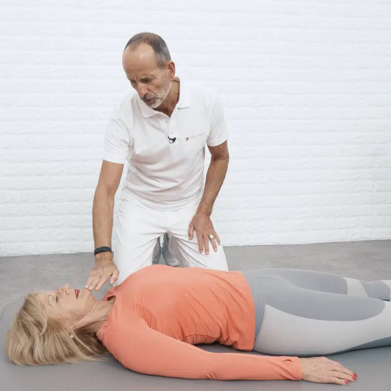 Roland Liebscher-Bracht kniet neben einer Frau, die gerade ausgestreckt am Boden liegt. Er berührt mit seiner Hand ihr Kinn und richtet ihren Kopf und Nacken gerade aus.