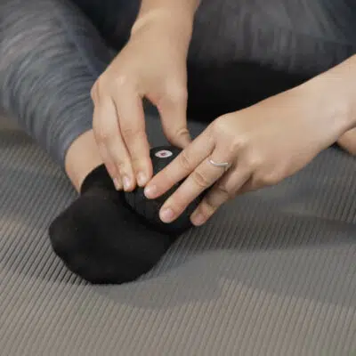 Eine Frau hält in ihren Fingerspitzen eine Mini-Faszienkugel von Liebscher & Bracht. Der Fuß dieser Frau liegt seitlich auf dem Boden und sie rollt mit der Kugel über ihre Fußsohle.