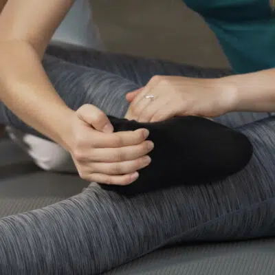 Eine Frau hat ihren rechten Fuß auf ihr linkes Knie gelegt. Mit ihrer rechten Hand hat sie ihre Zehen und den Vorfuß gegriffen und zieht sie für eine Dehnübung nach oben.