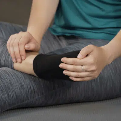 Eine Frau hat ihren rechten Fuß auf ihr linkes Knie gelegt. Mit ihrer rechten Hand hält sie den Knöchel fest, mit ihrer linken Hand zieht sie die Zehen und den Vorfuß für eine Dehnübung nach unten.