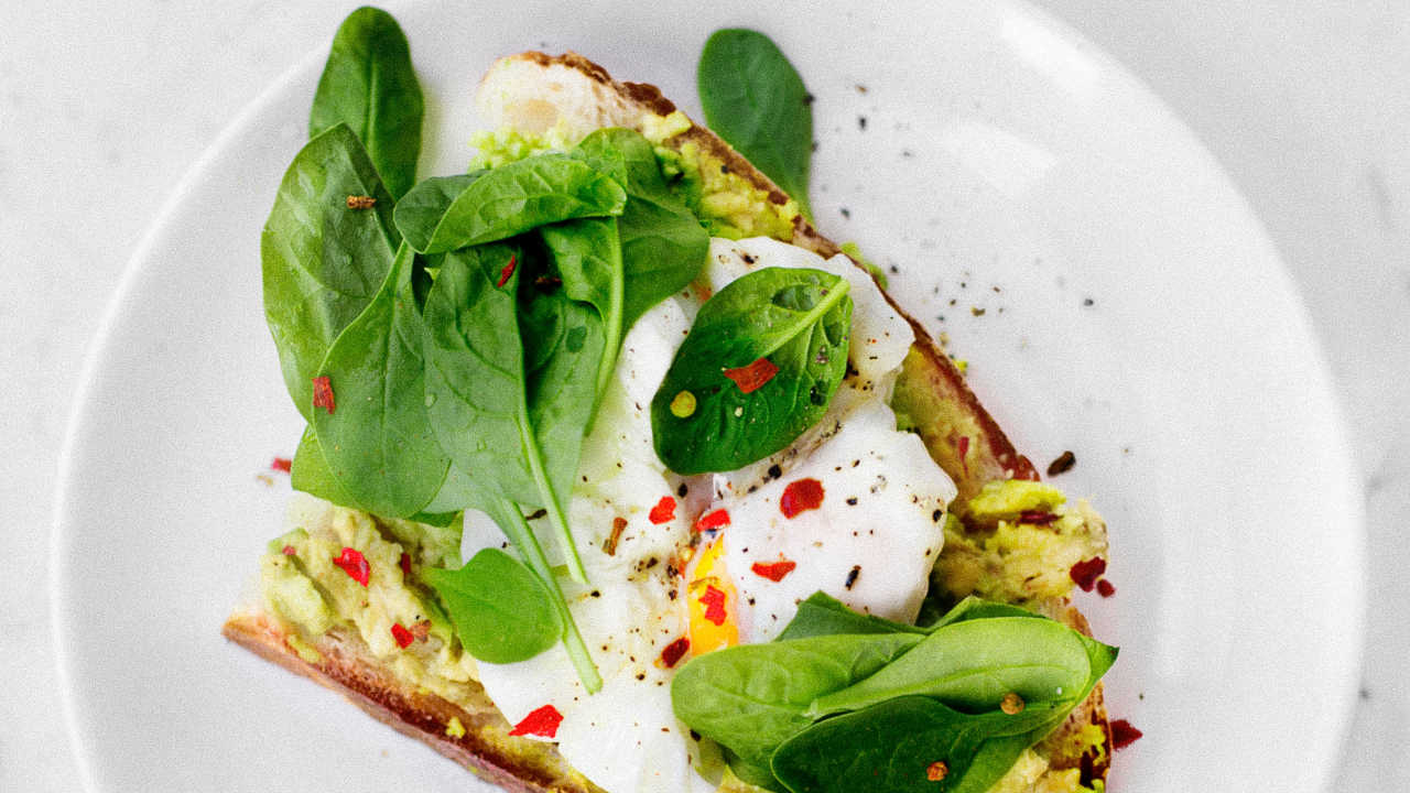 Eine Scheibe Sauerteigbrot mit Avocadoaufstrich, einem pochierten Ei und frischem Basilikum liegt auf einem weißen Teller