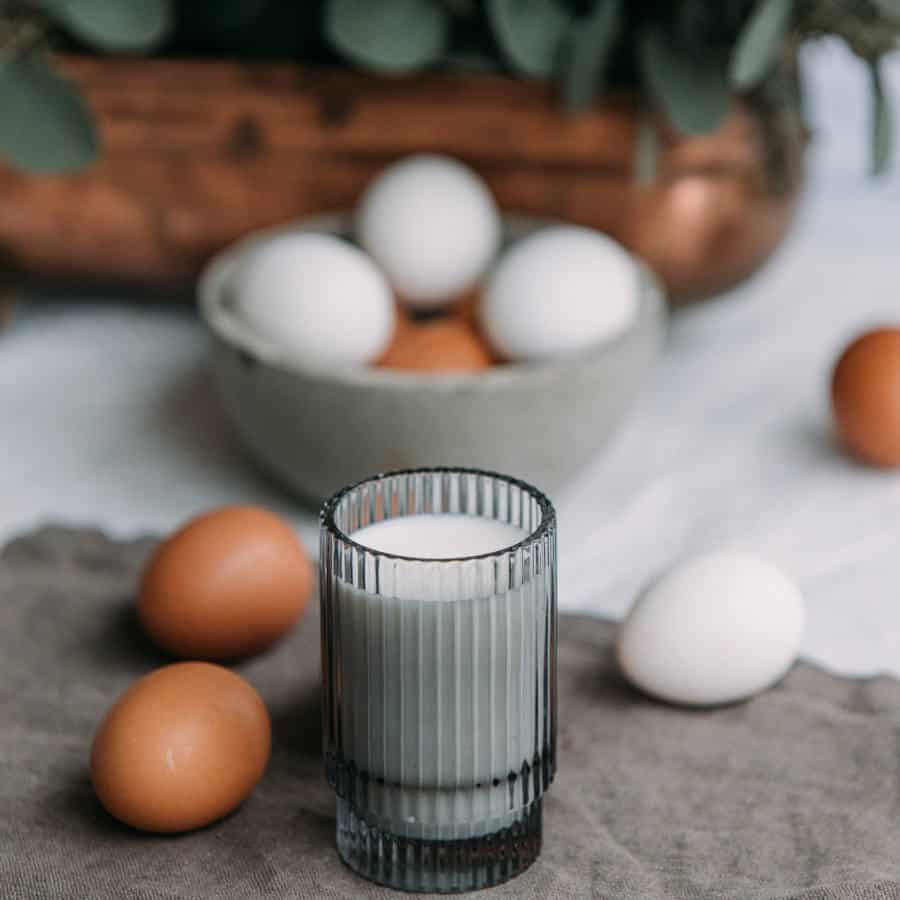 Ein Glas Milch steht im Vordergrund des Bildes. Dahinter liegen braune und weiße Eier auf dem Tisch verteilt und in einer Schüssel. Hinter der Schüssel ist ein Pflanzenkübel ansatzweise zu sehen.