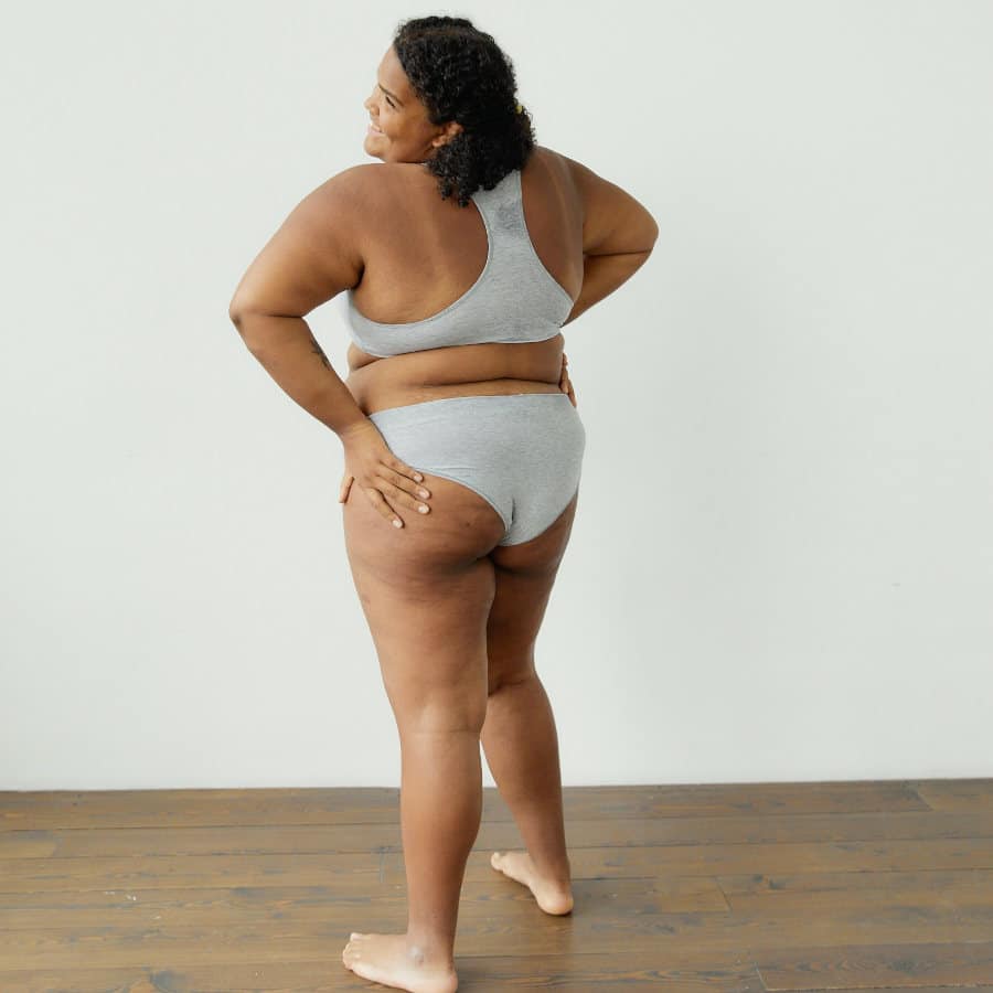 Eine junge, etwas fülligere Frau mit kurzen dunklen Haaren steht mit dem Rücken zur Kamera. Ihr Kopf ist zur linken Seite geneigt und ihre Arme in ihre Hüften gestemmt.