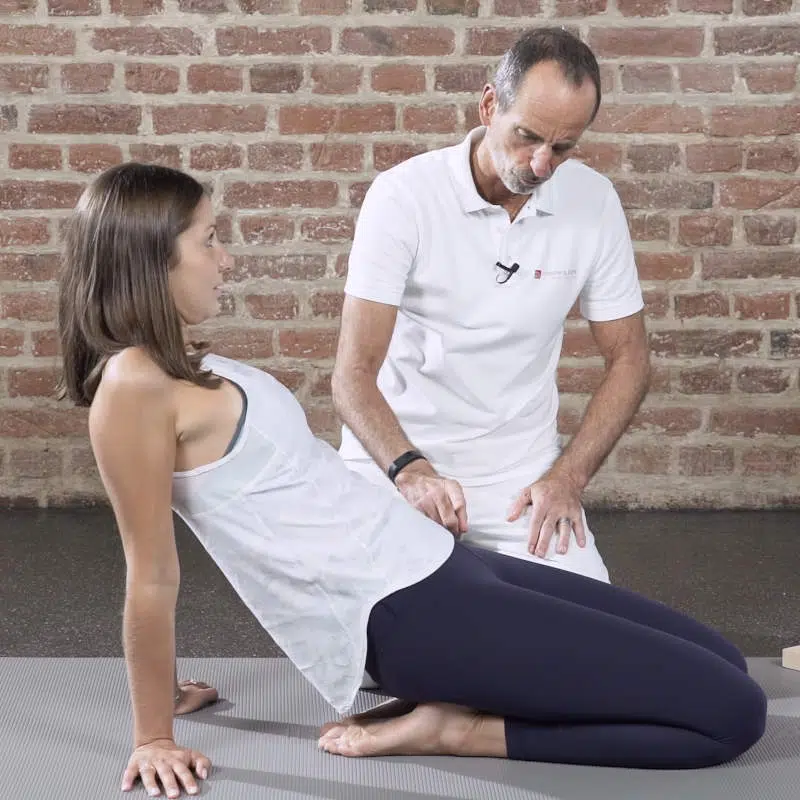 Roland Liebscher-Bracht kniet neben einer Frau und erklärt ihr eine Dehnübung gegen Knieschmerzen. Die junge Frau sitzt auf ihren Fersen und hat ihren Oberkörper nach hinten geneigt.
