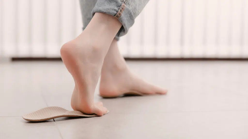 Eine Person steht barfuß auf zwei Einlegesohlen für die Schuhe. Der vordere Fuß liegt auf, während der hintere Fuß zum Gehen angehoben ist, sodass man die Fußsohle und die Ferse sieht.