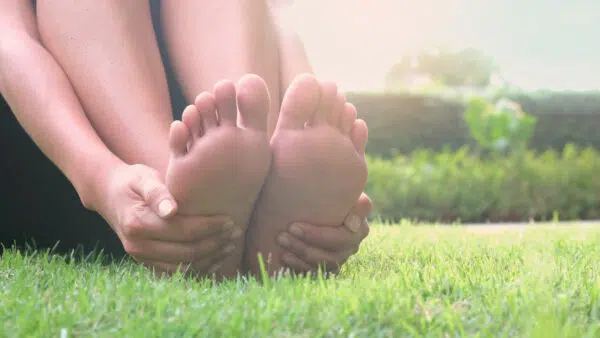 Eine Person sitzt barfuß im Gras und fasst sich mit beiden Händen an ihre Fußsohlen.