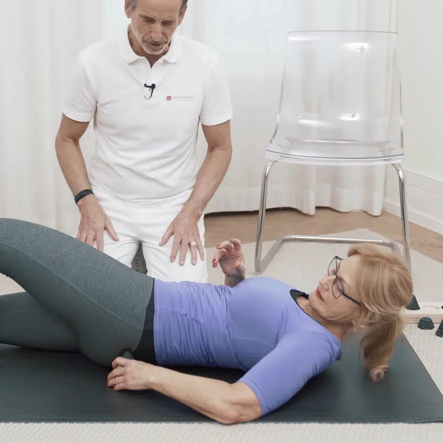 Eine Person liegt in Rückenlage am Boden und platziert den Drücker unter ihrem Gesäß.