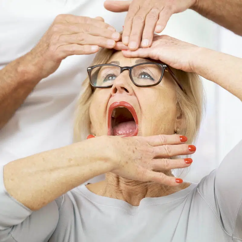 Eine Frau macht eine Übung zur Entspannung ihrer Kaumuskulatur. Dazu hat sie ihren Mund weit geöffnet. Sie hält ihren Unterkiefer mit einer Hand fest, während sie die andere Hand auf ihrer Stirn abgelegt hat.