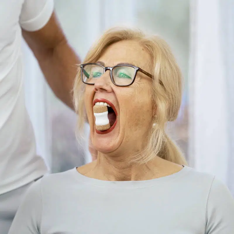 Eine Frau dehnt ihre Kiefermuskulatur mit dem Kieferretter von Liebscher & Bracht. Das Hilfsmittel ist zwischen ihren vorderen Schneidezähnen und sie hat ihren Mund weit geöffnet.