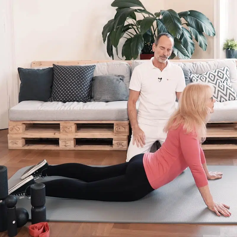 Roland Liebscher-Bracht erklärt einer Frau eine Übung gegen die Schambeinentzündung. Die Frau ist am Boden auf einer Trainingsmatte, ihre Beine sind ausgestreckt und sie drückt mit den Armen ihren Oberkörper nach oben.