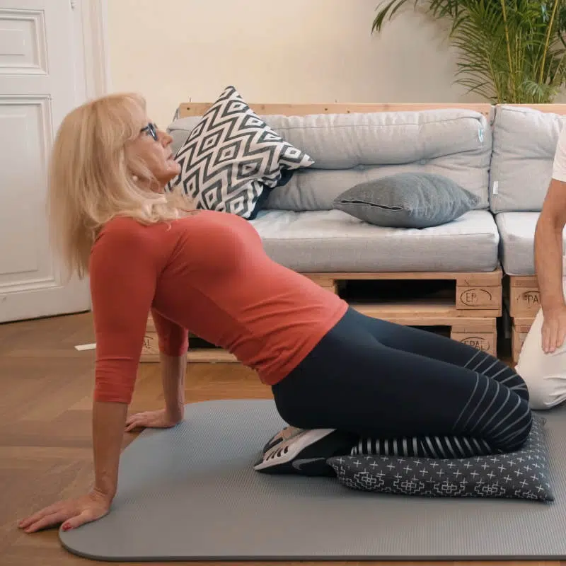 Eine Frau kniet am Boden auf einem Kissen. Sie sitzt dabei vollständig auf ihren Fersen. Sie neigt ihren Oberkörper bei dieser Übung nach hinten und stützt sich mit den Händen hinter ihrem Rücken ab.