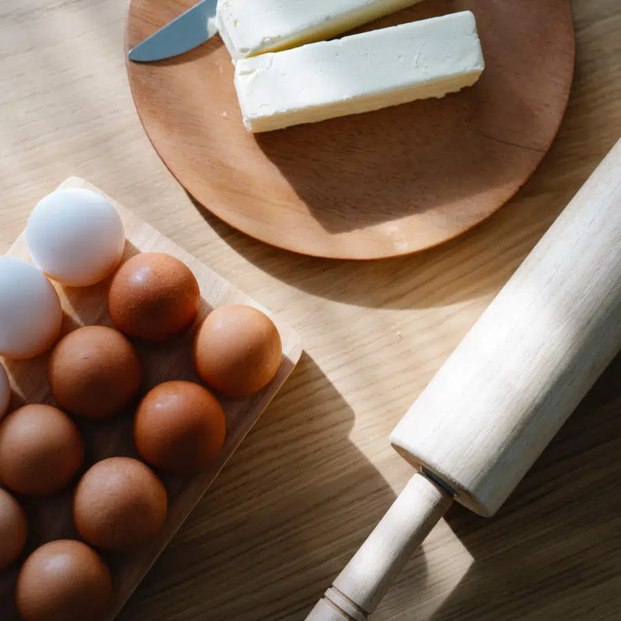 Zwei Stücke Margarine liegen auf einem Holzbrett. Daneben liegt ein Brotmesser. Links unten sind braune und weiße Eier zu sehen. Daneben liegt ein Nudelholz, um Teig auszurollen.