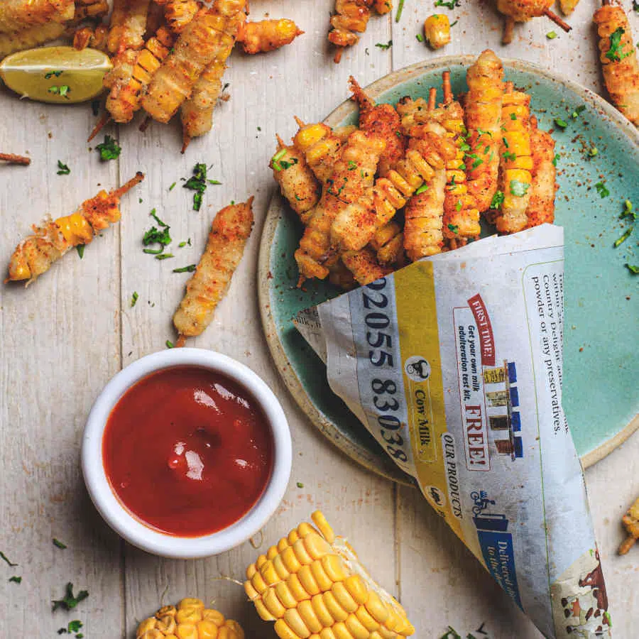 Eine Tüte mit frittierten Pommes aus Mais liegt auf einem blauen Teller. Auf dem Tisch sind weitere Pommes und ein Schälchen Ketchup zu sehen.