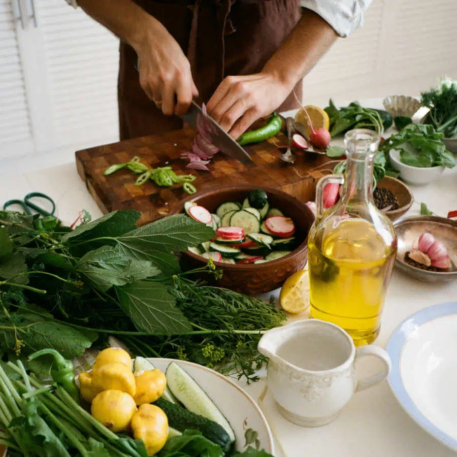 Eine Person schneidet Zutaten für einen bunten Salat klein. Salat, frische Kräuter, Radieschen, Lauch und anderes Gemüse liegen auf dem Tisch vor ihr. Eine große Flasche mit Pflanzenöl steht auch dabei.