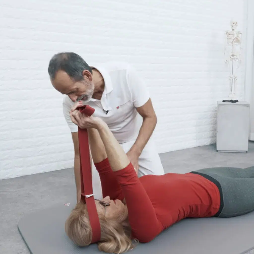 Eine Person liegt am Boden in Rückenlage und hat die Übungsschlaufe um ihren Kopf gelegt. Ihre Arme sind nach oben gestreckt und sie hält das andere Ende der Schlaufe in den Händen, sodass die Schlaufe auf Spannung ist. Der Kopf schwebt ein paar Zentimeter über dem Boden.