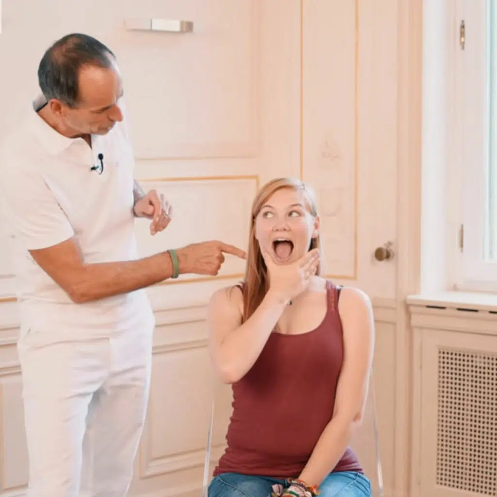 Eine Person sitzt auf einem Stuhl und hat die rechte Hand am Unterkiefer. Damit zieht sie den Unterkiefer nach unten, sodass der Mund geöffnet ist.