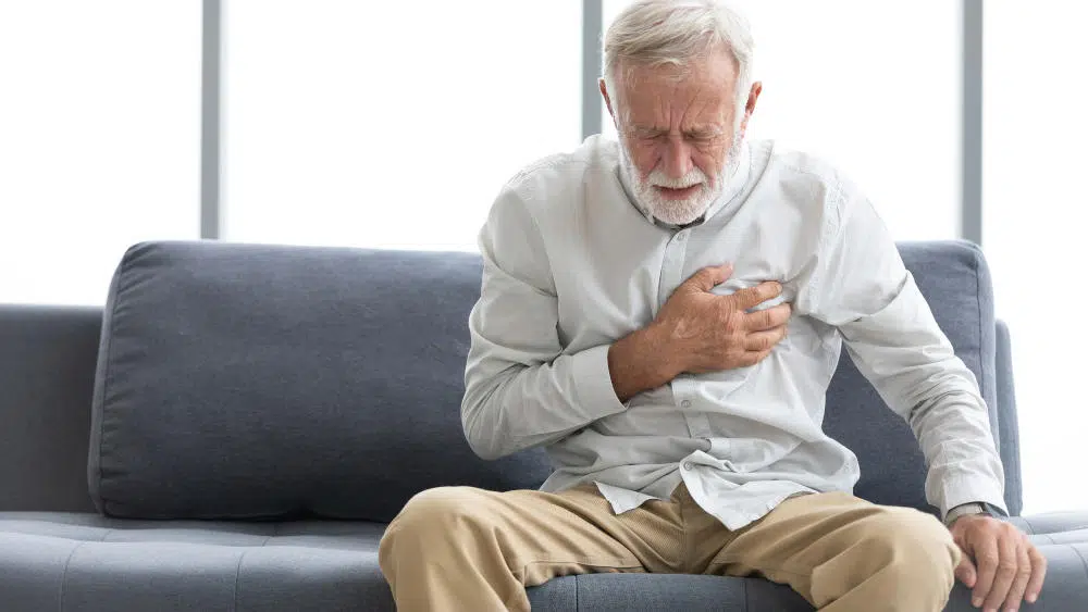 Ein älterer Herr sitzt auf dem Sofa und fasst sich mit schmerzerfülltem Gesicht an sein Herz.