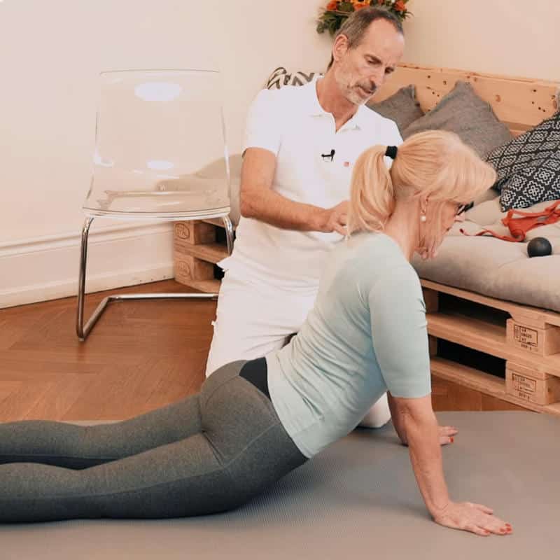 Roland Liebscher-Bracht zeigt einer Frau eine Dehnübung. Die Frau liegt mit ihren Beinen am Boden und stützt ihren Oberkörper mit gestreckten Armen vom Boden weg. Roland Liebscher-Bracht korrigiert dabei die Position ihrer Halswirbelsäule.