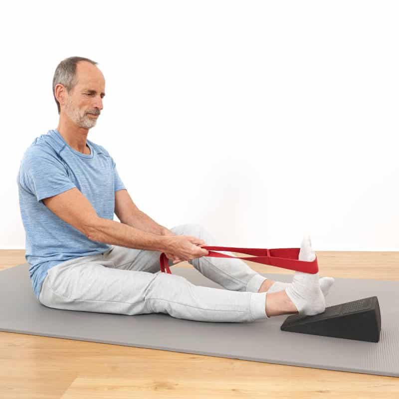 Roland Liebscher-Bracht sitzt mit ausgestreckten Beinen am Boden. Um die Zehen an einem Fuß ist eine rote Schlaufe geschwungen, an der er den Fuß zu sich zieht.