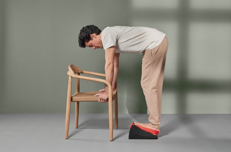 Ein junger Mann in weißem T-Shirt und beiger Baumwoll-Hose steht mit seinem linken Bein auf dem Knieretter. Der Fuß steht in der roten Trittschlaufe, damit er nicht abrutscht. Er beugt sich mit geradem Rücken nach unten, um sich an den Seiten der Sitzfläche eines Holzstuhls abzustützen.