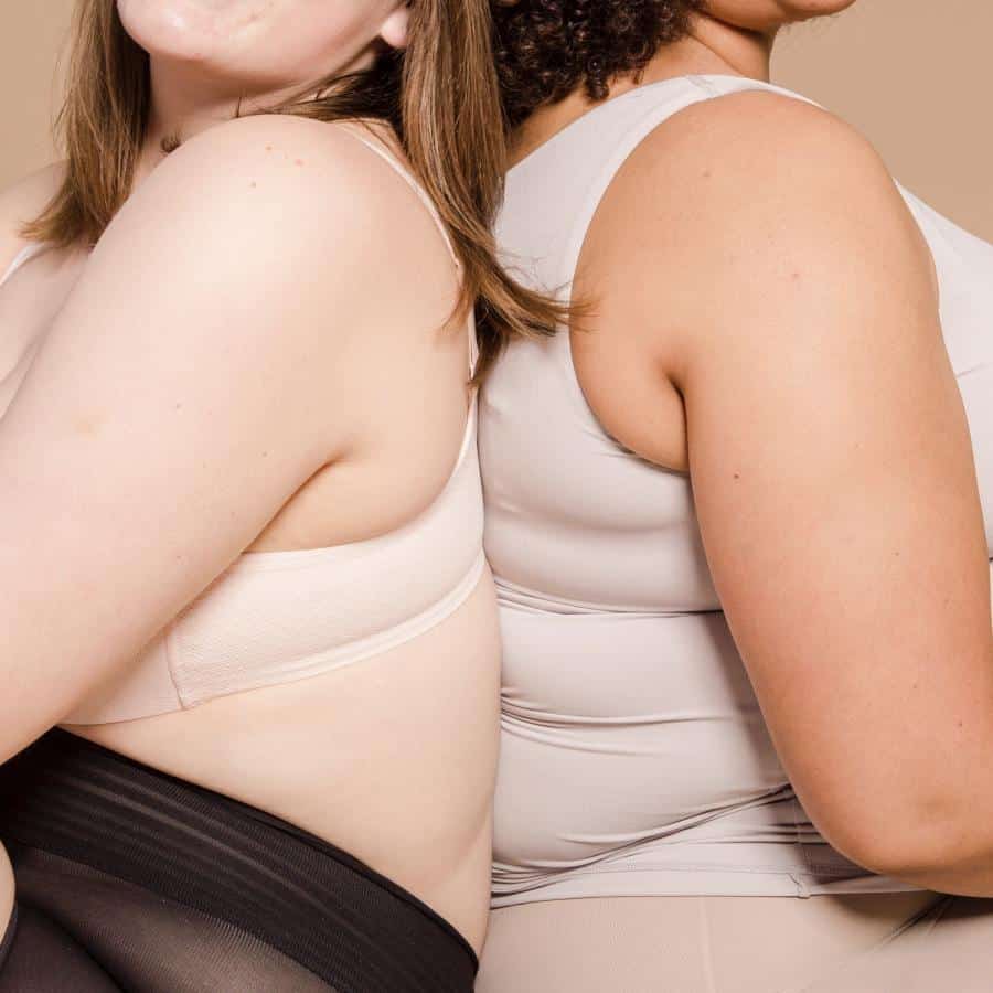 Zwei Frauen sitzen in Unterwäsche Rücken an Rücken. Ihre Köpfe sind der Kamera zugewandt, wobei von ihrem Gesicht nur der Kiefer und auch ihre Haare zu sehen sind.