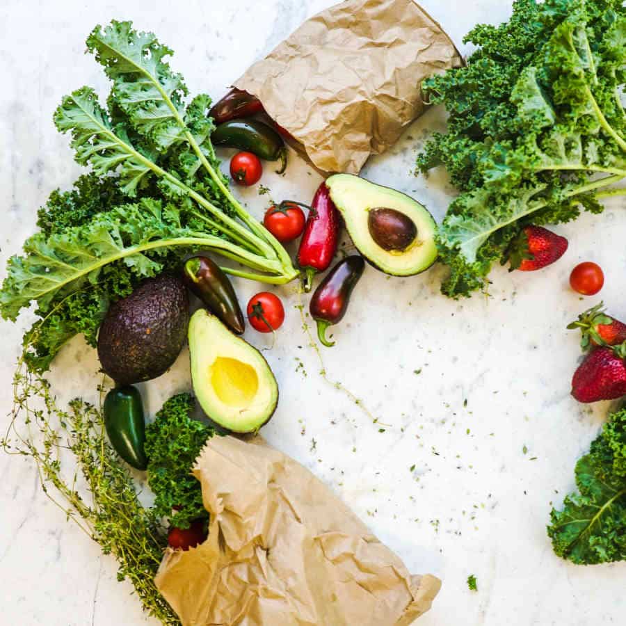 Verschiedenes Gemüse wie Avocado, Grünkohl, Spitzpaprika und auch ein paar Erdbeeren liegen auf einer Marmorplatte verteilt. Zwei braune Markttüten, in denen das Gemüse war, liegen zwischen den pflanzlichen Nahrungsmitteln.