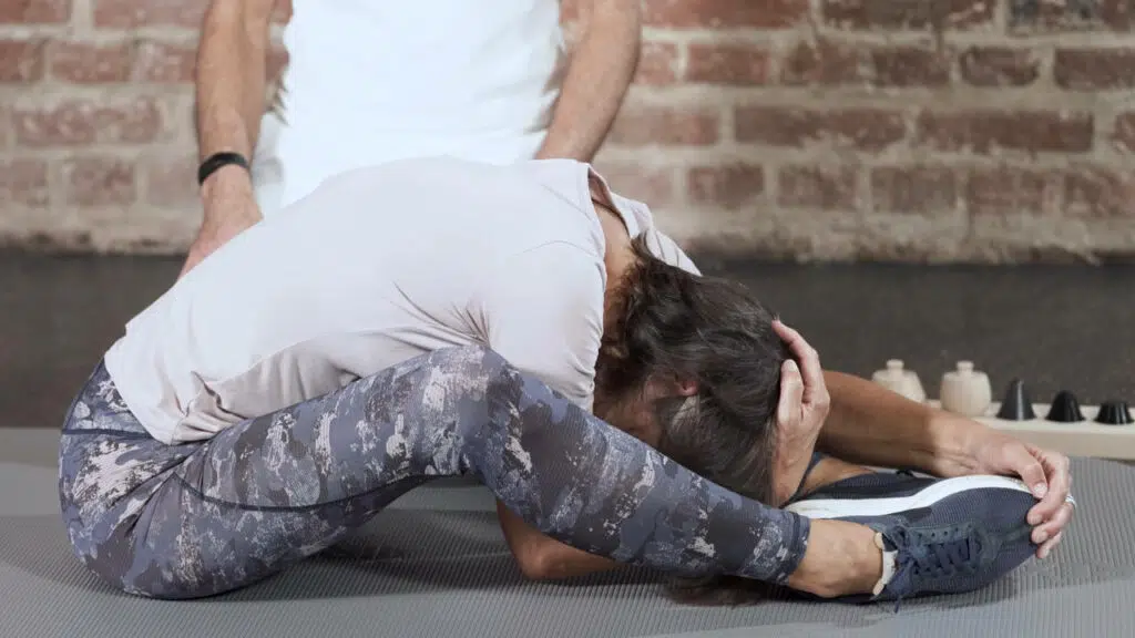 Eine Frau sitzt auf einer Übungsmatte. Ihre Beine lässt sie nach links und rechts fallen und ihren Oberkörper beugt sie nach vorne.