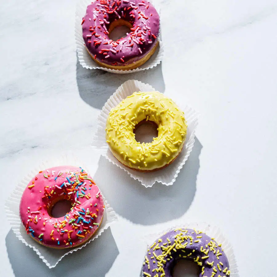 Es sind vier Donuts zu sehen, die mit verschieden bunten Glasuren und Streuseln dekoriert sind. Unter ihnen liegt jeweils eine Art Papier zum in die Hand nehmen.