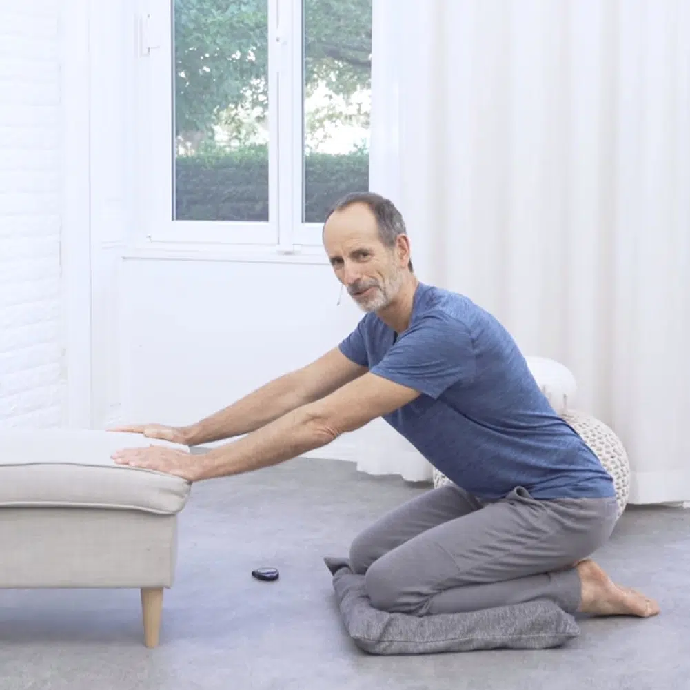 Eine Person sitzt vor einem Hocker auf dem Boden im Fersensitz. Die Knie sind durch ein Kissen gepolstert. Beide Hände hat die Person auf den Hocker abgelegt.