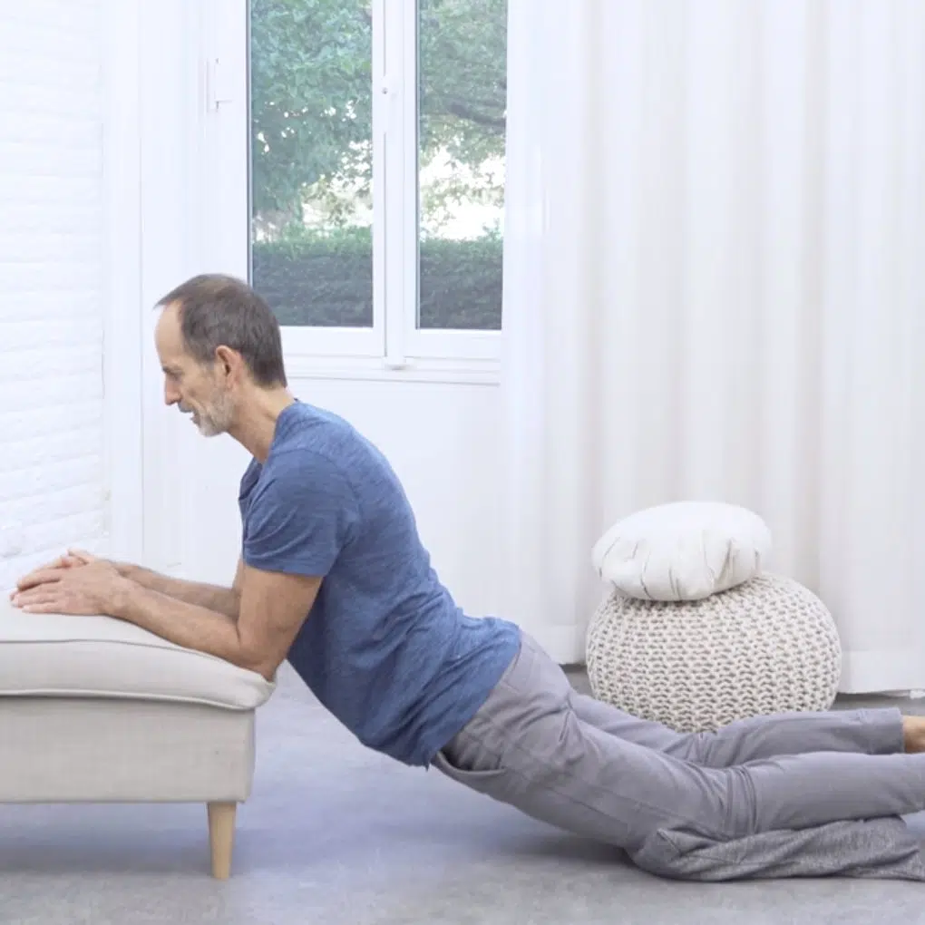 Eine Person hat beide Unterarme auf einem Hocker abgelegt. Sie hat ihre Knie am Boden durch ein Kissen gepolstert und lässt das Becken nach unten sinken, sodass der gesamte Körper in eine Rückbeuge kommt.