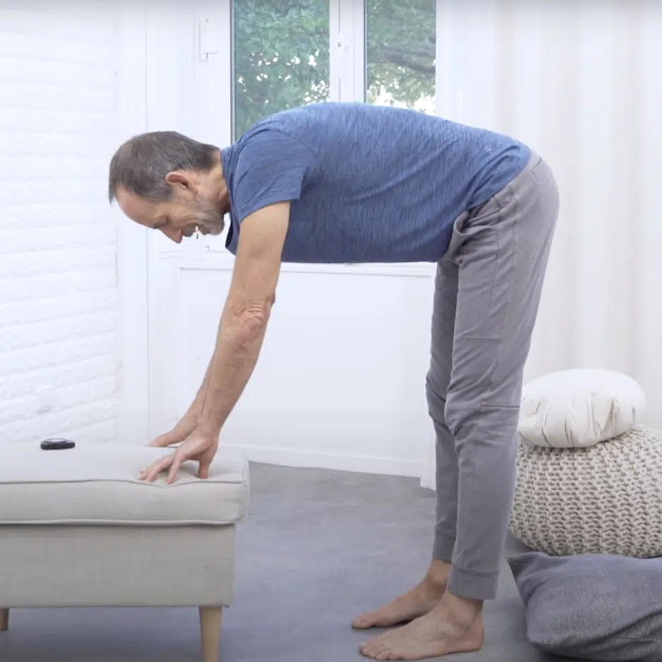 Eine Person steht mit ausgestreckten Beinen vor einem Hocker und beugt sich nach vorne. Beide Hände sind auf dem Hocker abgelegt und sie lässt sich weiter nach unten sinken.