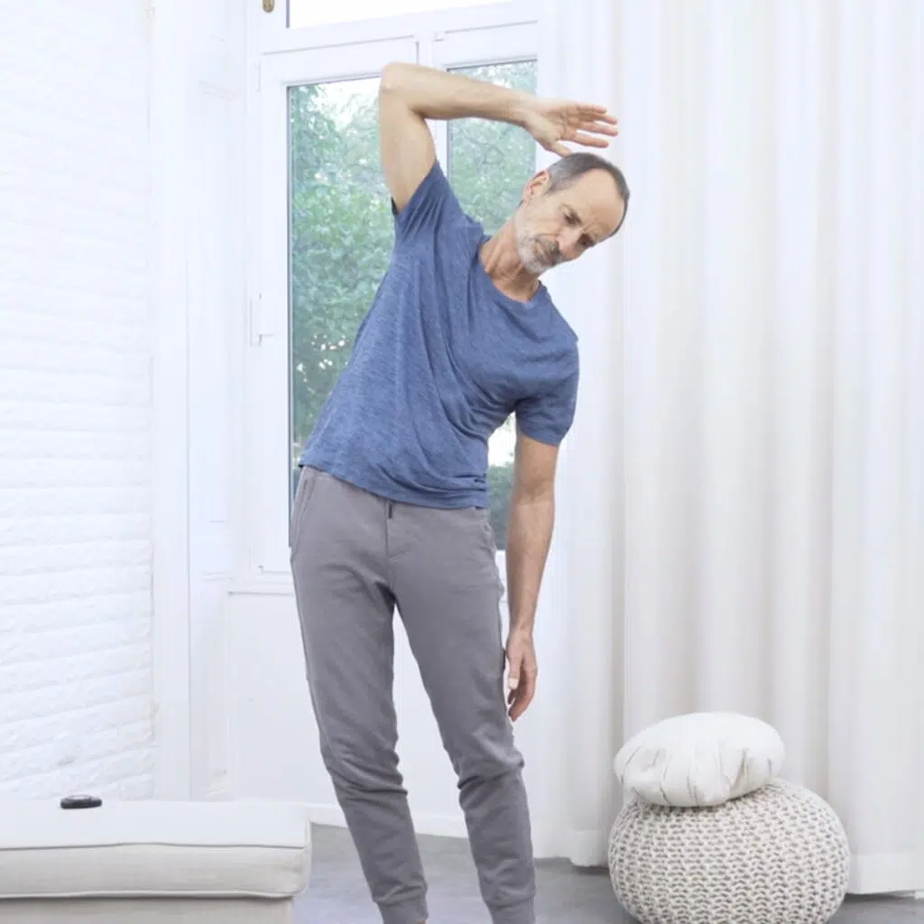 Eine Person steht mit ausgestreckten Beinen auf dem Boden. Sie neigt ihren Oberkörper nach links und hebt den rechten Arm nach oben. Der rechte Arm ist angewinkelt, sodass der Ellbogen nach oben zeigt.