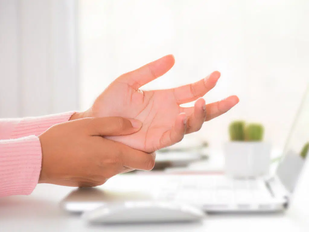 Eine rechte Hand umfasst das Handgelenk der linken, nach oben geöffneten Hand vor einem Laptop.