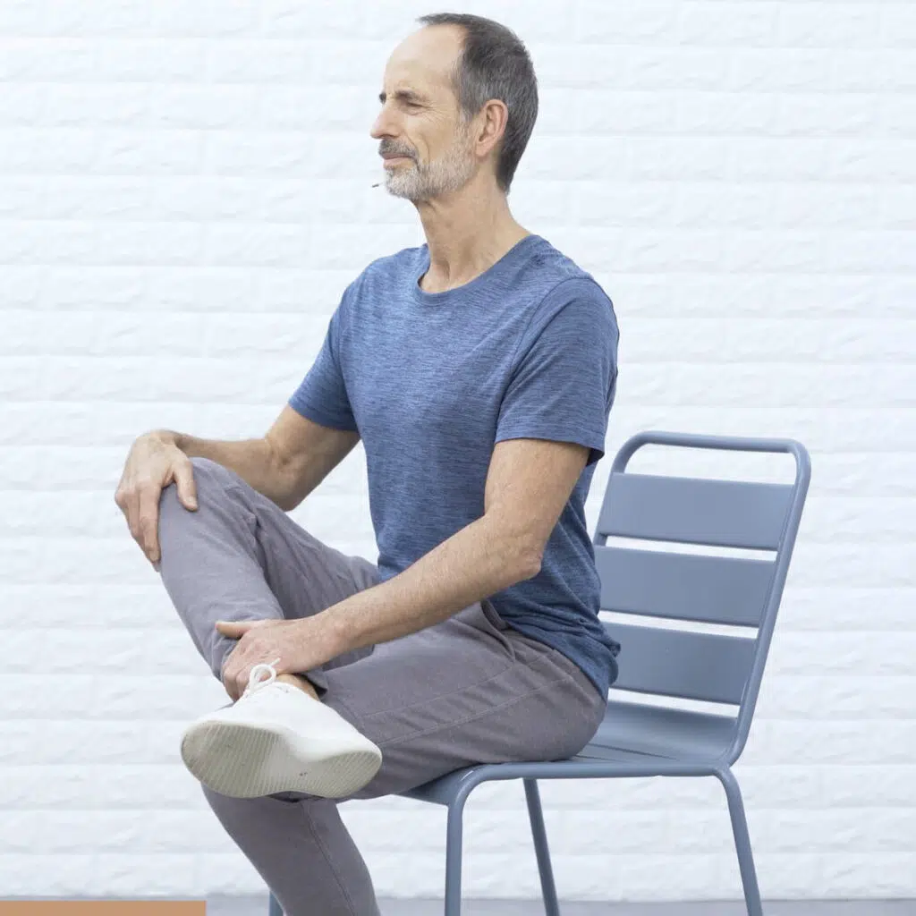 Roland Liebscher-Bracht sitzt auf einem Stuhl, hat sein rechtes Bein auf sein linkes gelegt und beugt sich mit geradem Rücken nach vorne.