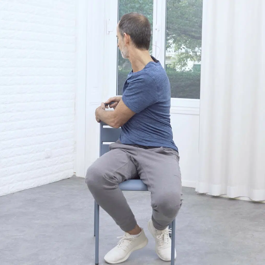 Roland Liebscher-Bracht sitzt auf einem Stuhl und dreht sich nach rechts. Dafür hält er sich an der Stuhllehne fest.