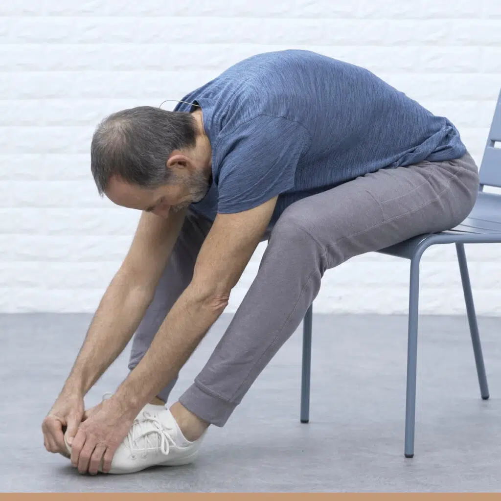 Roland Liebscher-Bracht sitzt auf einem Stuhl und beugt sich nach vorne, um nach seinen Füßen zu greifen.