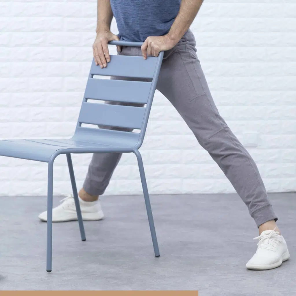 Roland Liebscher-Bracht steht mit weitgespreizten Beinen hinter einem Stuhl. Er hält sich an der Lehne fest.