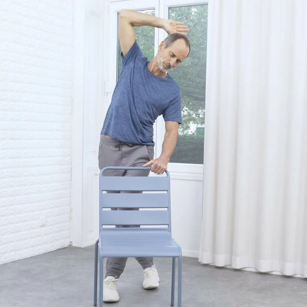 Roland Liebscher-Bracht steht hinter einem Stuhl und beugt sich weit zur linken Seite. Sein langgestreckter rechter Arm über seinem Kopf zieht ihn.