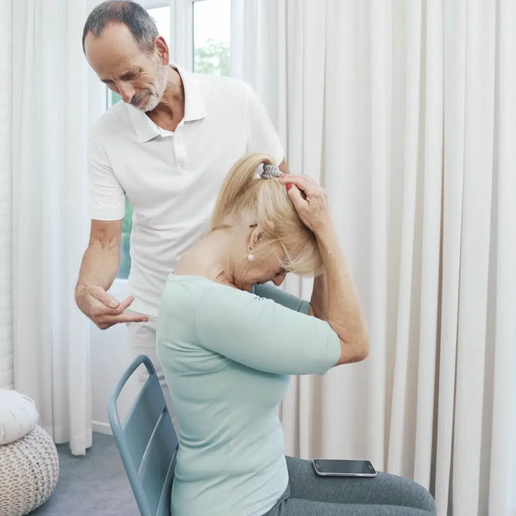 Roland Liebscher-Bracht steht vor einer Patientin und zeigt eine Nackenübung. Er deutet auf die Wirbelsäule. Die Patientin sitzt gerade auf einem Stuhl und hat beide Hände an den Kopf gelegt. Sie zieht den Kopf nach vorne, um die Nackenmuskulatur zu dehnen.