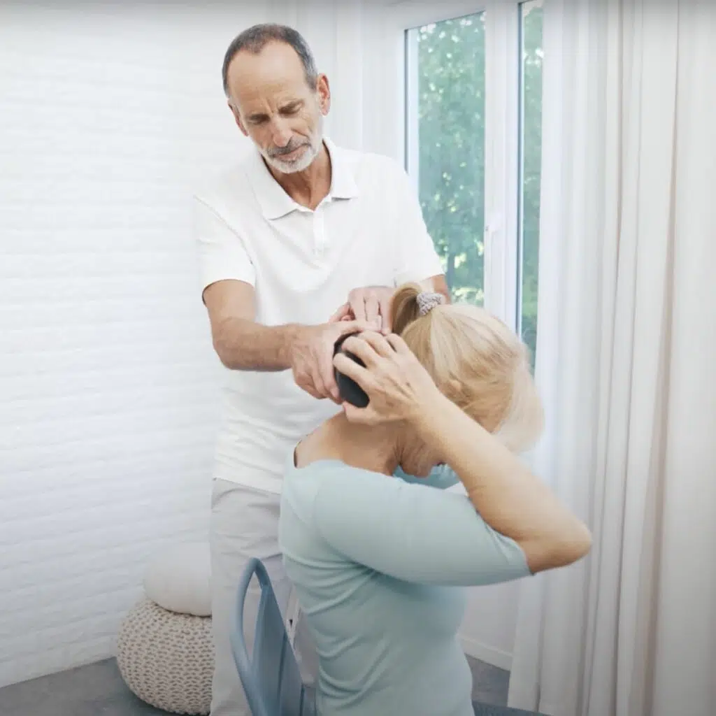 Roland Liebscher-Bracht hält eine Faszienrolle an den Nacken einer Patientin. Sie umgreift die Rolle mit beiden Händen und rollt an der Halswirbelsäule hinab.