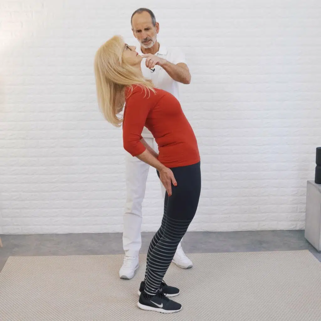 Eine Frau steht hüftbreit, ihre Arme sind oberhalb ihres Gesäßes aufgestemmt während sie sich mit dem Oberkörper weit nach hinten lehnt.