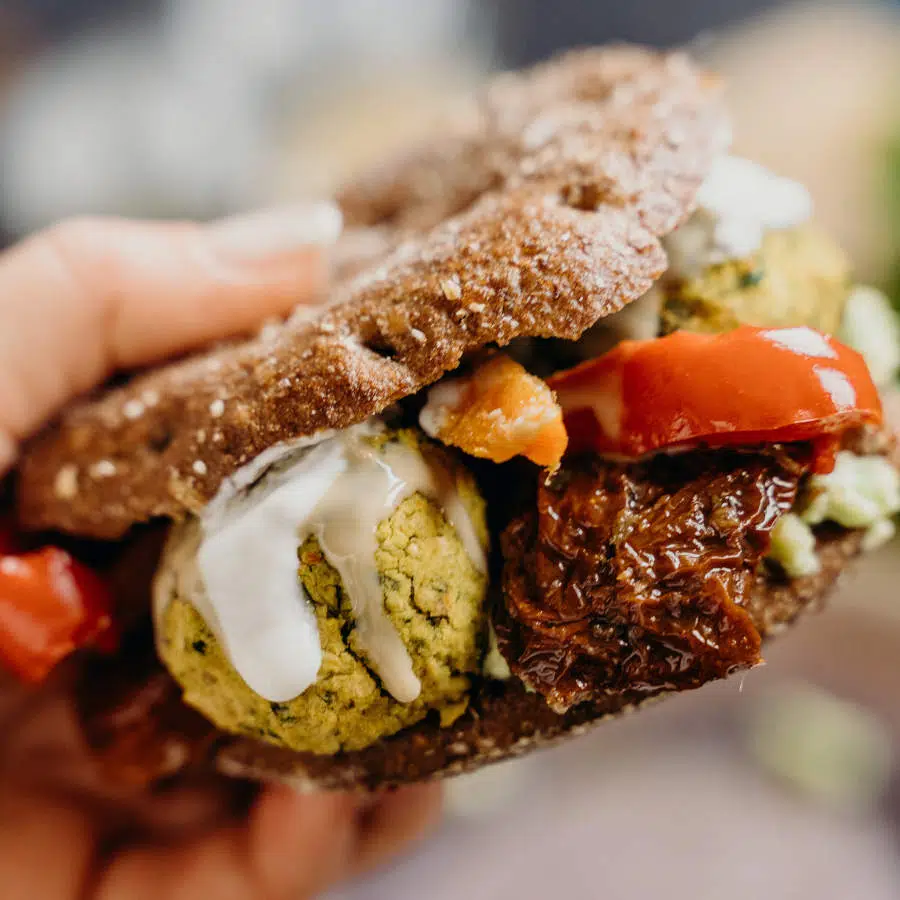 Eine Hand hält einen selbstgemachten Falafel-Burger mit Vollkornbrötchen und Tomaten in die Kamera.