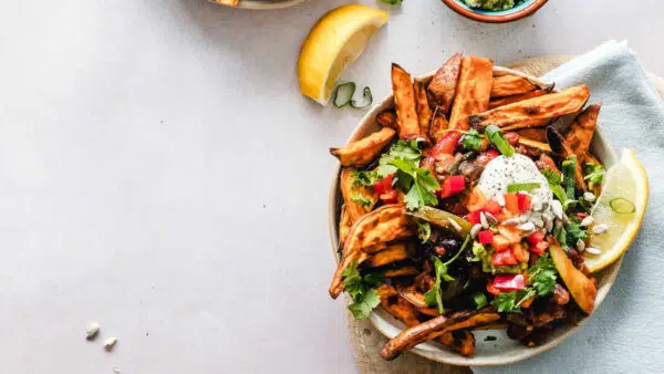Ein großer Teller mit selbstgemachtes Süßkartoffelpommes und Gemüse steht auf einem weißen Tisch. Links oben liegt ein Stück Zitrone.