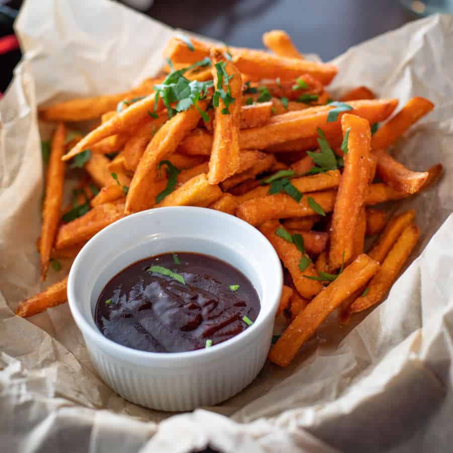 Süßkartoffelpommes liegen auf einem Teller, der mit Papier ausgekleidet ist. Daneben steht ein Schälchen mit Ketchup.